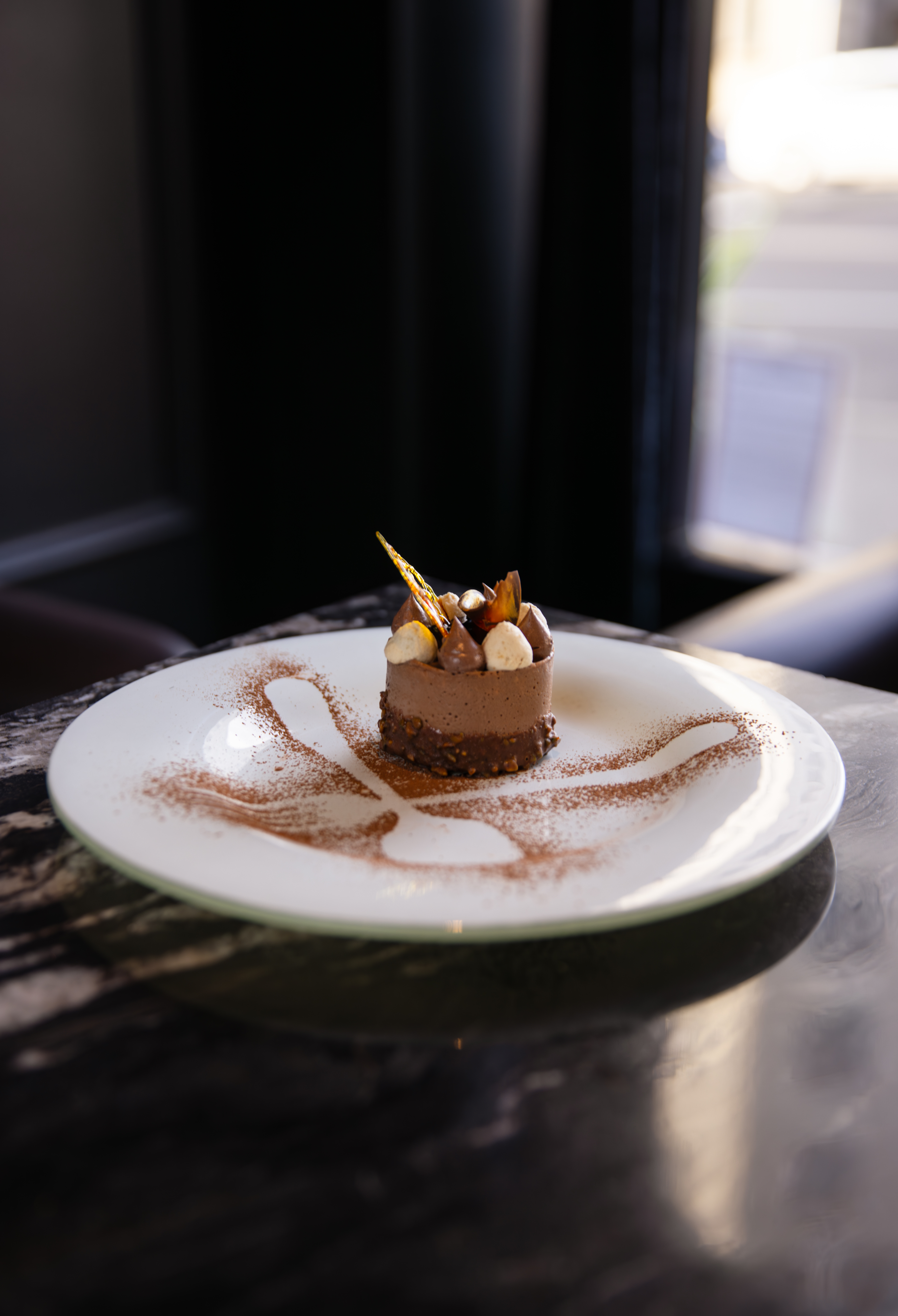 Individual chocolate mousse cake topped with truffles, marshmallows, and a gold chocolate shard, plated with an artistic cocoa powder swirl.