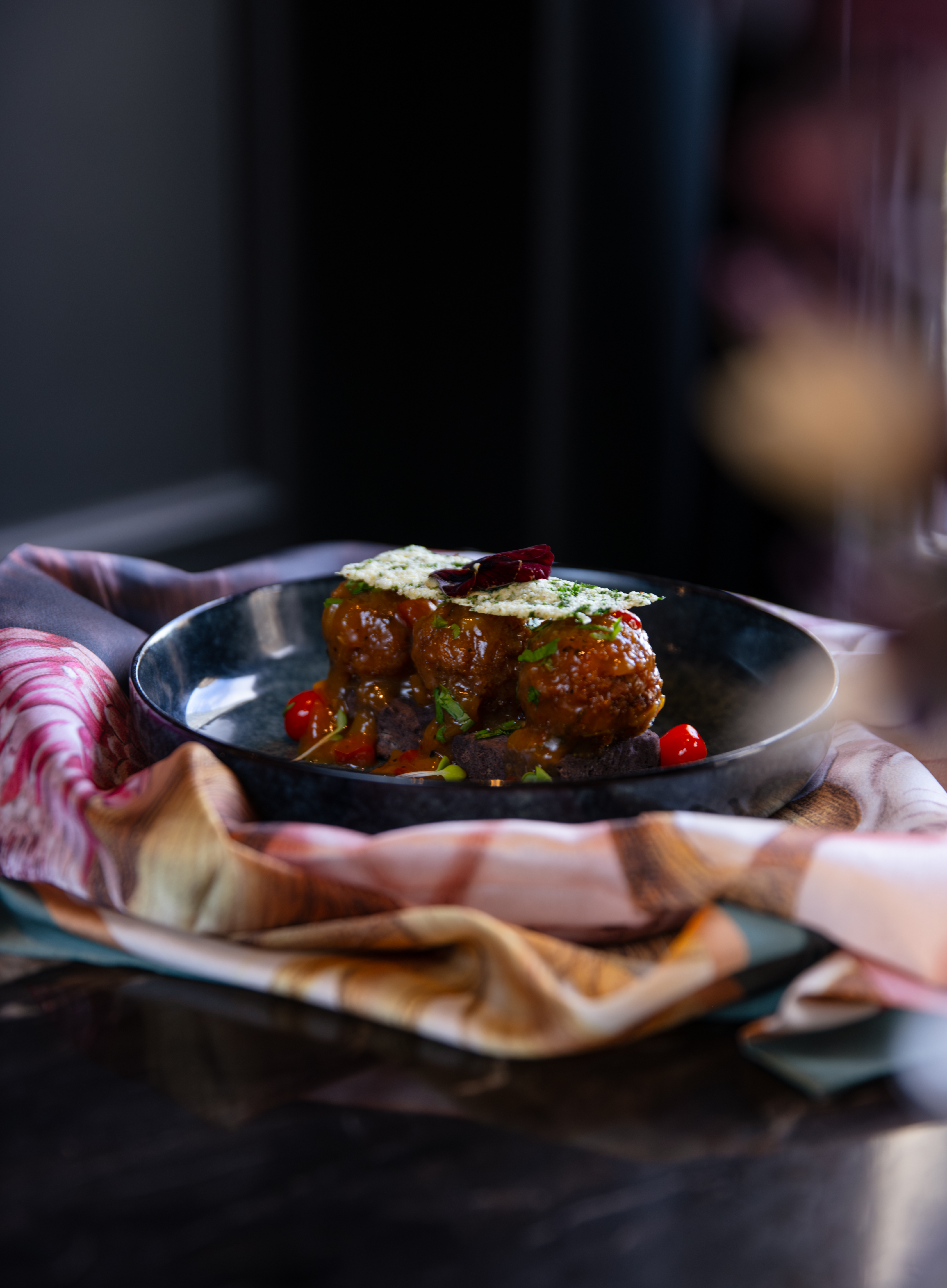 Crumbed meatballs in tomato sauce topped with a herb parmesan crisp, served in a dark ceramic bowl on a decorative napkin."