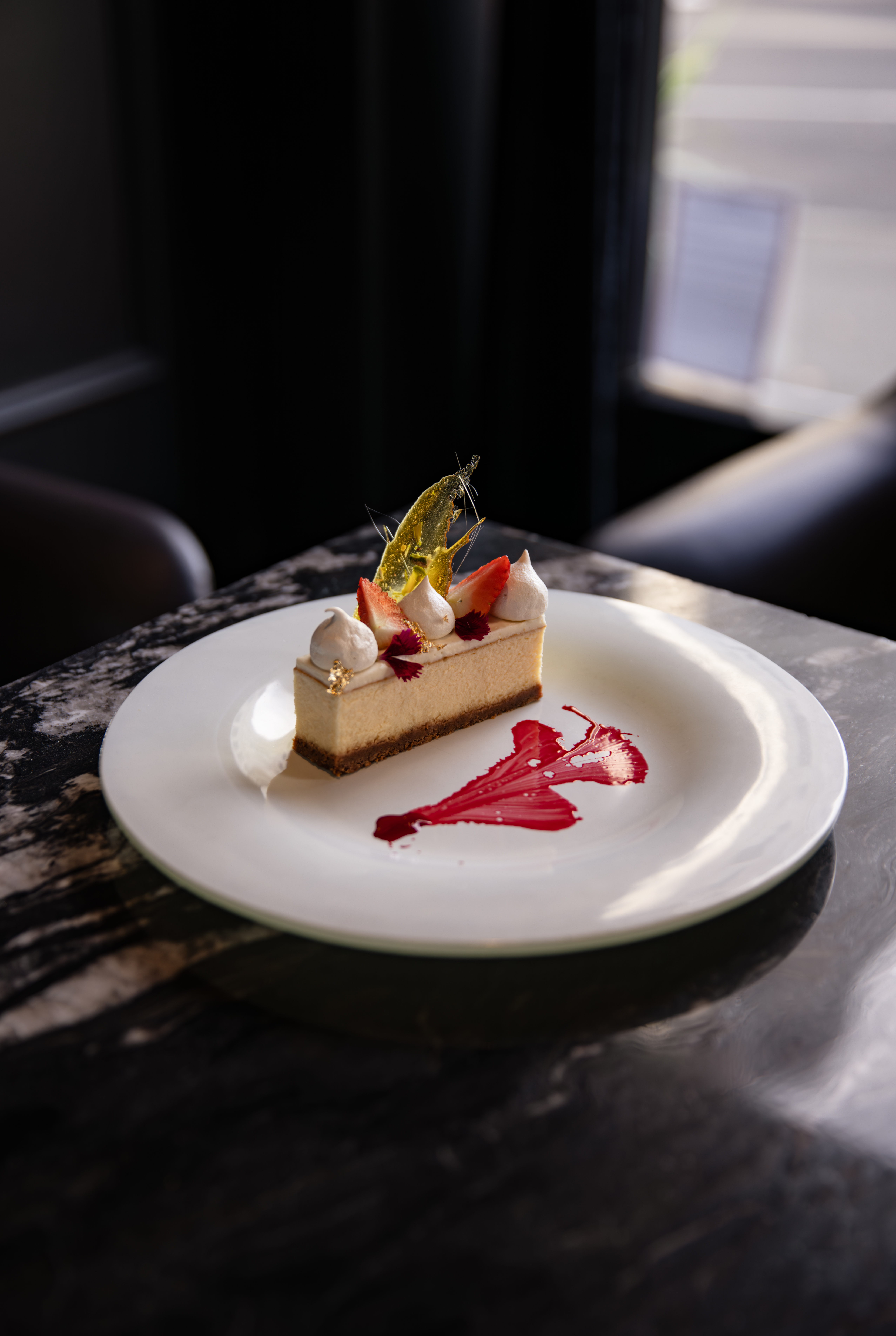 Cheesecake slice topped with meringue kisses, strawberries, and a gold sugar shard, with a raspberry coulis brushstroke on a white plate.