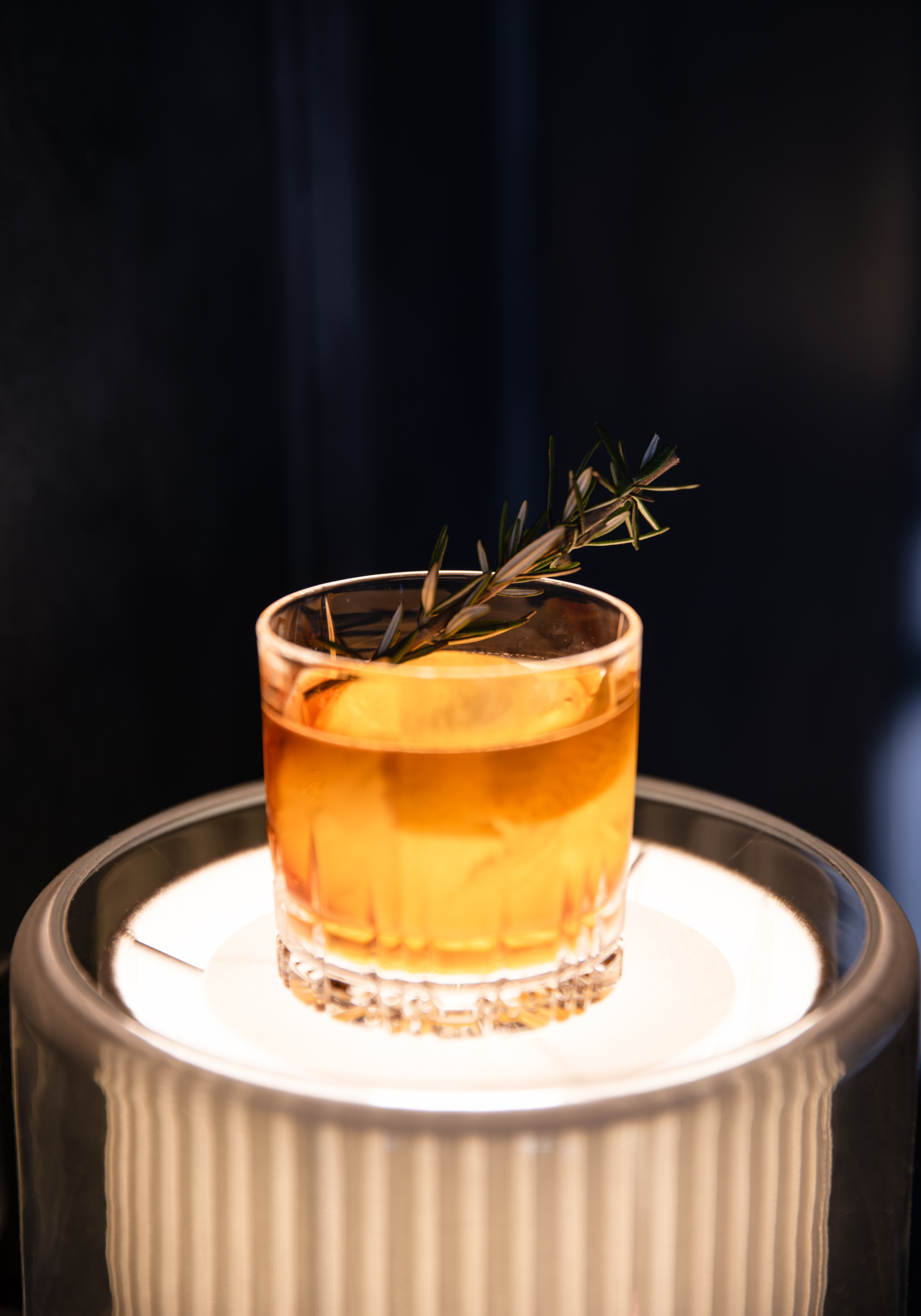 An amber whisky cocktail with an orange slice and fresh rosemary sprig garnish in a cut crystal rocks glass, glowing on an illuminated pedestal lamp against a dark background.